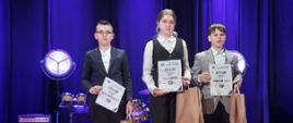 Troje uczniów stoi na scenie, każdy trzyma w ręce dyplom i torbę z upominkami