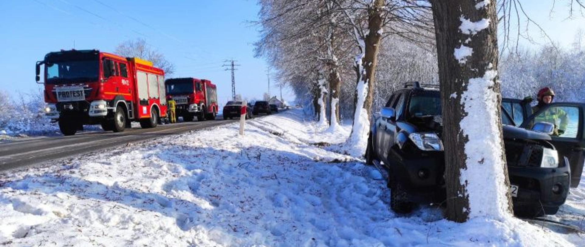 Centralny punkt zdjęcia stanowi rozbite auto terenowe. Auto koloru granatowego. Pojazd ma całkowicie uszkodzony przód. Po lewej na drodze pojazdy gaśnicze. Przy aucie stoi strażak.
