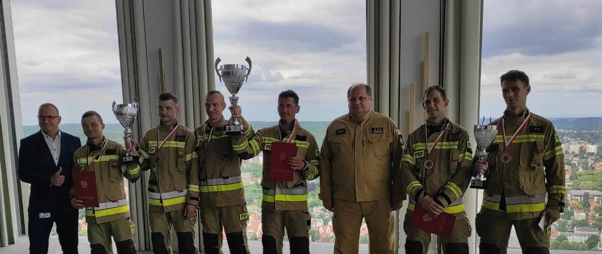 Zastępca pomorskiego komendanta wojewódzkiego Państwowej Straży Pożarnej oraz szef działu Zarządzania Bezpieczeństwem Olivia Business Centre stoją razem ze zwycięzcami biegu po schodach. Strażacy trzymają puchary oraz dyplomy.