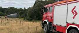 Na pierwszym planie stoi pojazd ratowniczo gaśniczy zapzarkowany na łące. W tle kolejny pojazd straży pożarnej, z którego podawana jest woda w kierunku zalesienia.