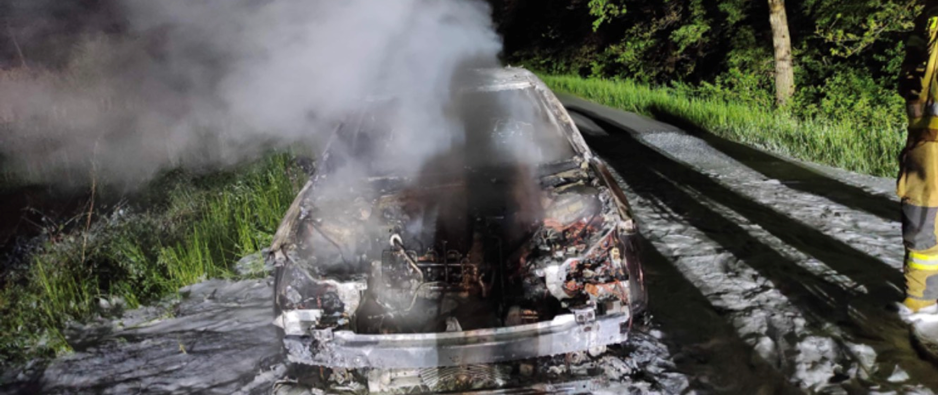 Zdjęcie przedstawia spalony samochód osobowy. Nad samochodem unosi się dym. Na jezdni pozostałości po pianie gaśniczej. Samochód całkowicie spalony