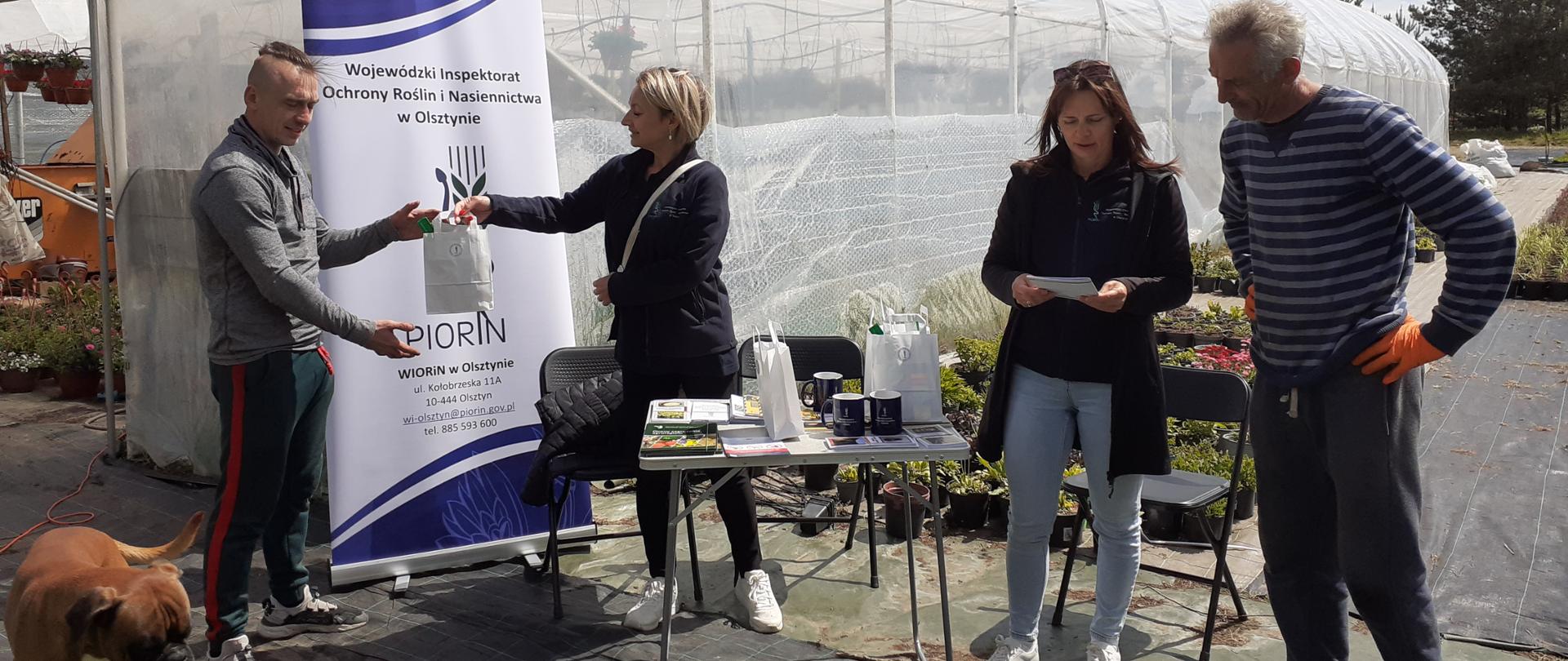 Grupa czterech osób stoi przy stoliku informacyjnym organizowanym przez WIORiN w Olsztynie
oddział w Piszu, wydarzenie odbywa się w na zewnątrz w Szkółce roślin ozdobnych, pogoda jest
słoneczna. W tle widać tunel foliowy i rośliny ozdobne. Jedna z inspektorek wręcza mężczyźnie
gadżet promocyjny, a kolejna omawia ulotkę informacyjną. Na stoliku leżą broszury i materiały
edukacyjne.