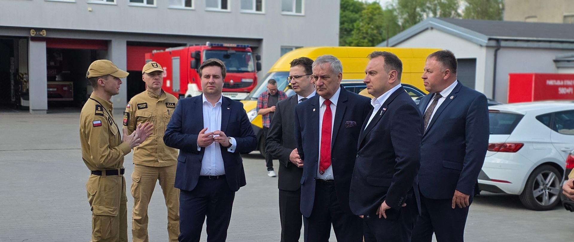 Na zdjęciu widać KP PSP w Zgierzu oprowadzającego gości - Wiceministra SWIA Wiesława Szczepańskiego wraz z Senatorem RP – Marcinem Karpińskim oraz Wicestarostą Powiatu Zgierskiego – Sebastianem Duninem po terenie jednostki , w tle czerwone bramy garażowe JRG w Zgierzu oraz samochód ratowniczo-gaśniczy. 