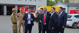 Na zdjęciu widać KP PSP w Zgierzu oprowadzającego gości - Wiceministra SWIA Wiesława Szczepańskiego wraz z Senatorem RP – Marcinem Karpińskim oraz Wicestarostą Powiatu Zgierskiego – Sebastianem Duninem po terenie jednostki , w tle czerwone bramy garażowe JRG w Zgierzu oraz samochód ratowniczo-gaśniczy. 