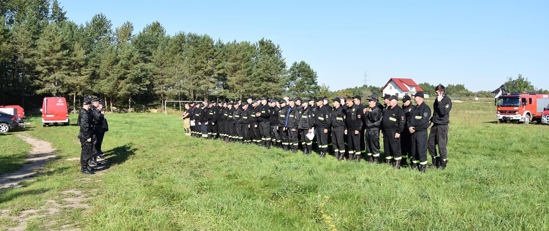 Zdjęcie obrazuje strażaków OSP i PSP ustawionych w szyku w tle samochody pożarnicze i budynek oraz las