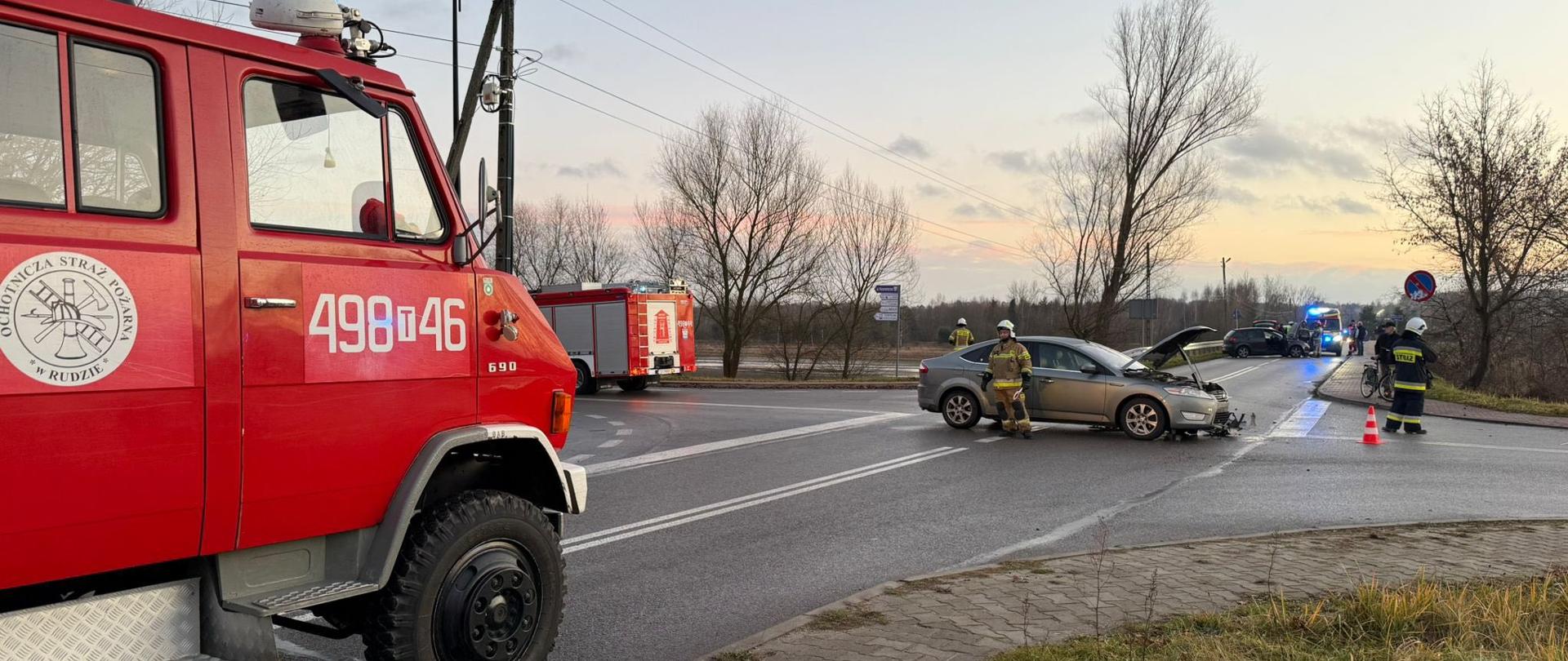 Skrzyżowanie ulic, na środku stoi uszkodzony pojazd osobowy obok strażacy i pojazdy strażackie