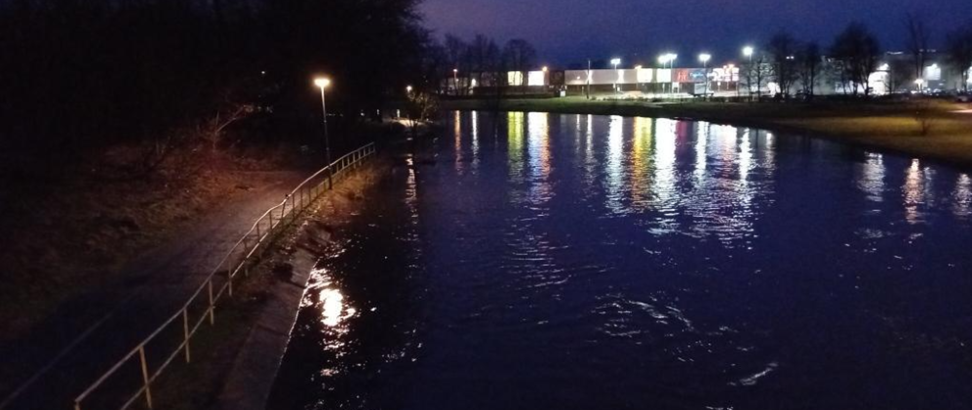 Na zdjęciu widać stan rzeki Gwdy na 12.02.2024 na godzinę 6:50. Jest jeszcze ciemno choć już świta. Widać światła i budynki miasta Piły. Stan rzeki wysoki. Po lewej deptak, drzewa i barierki. 