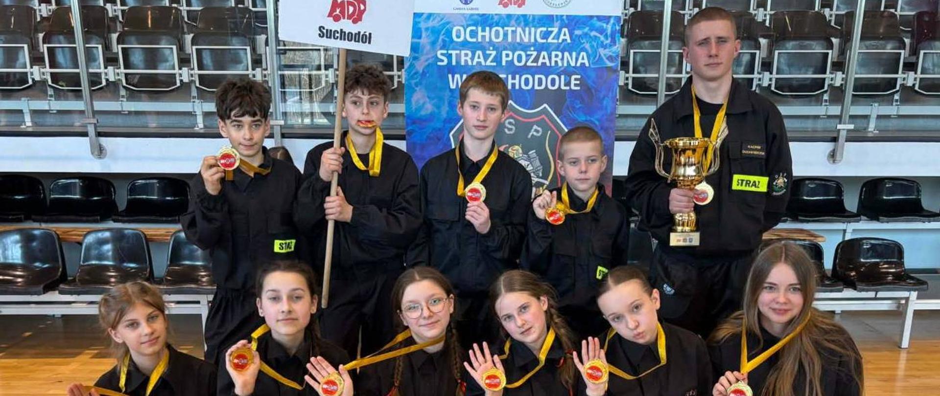 Zdjęcie przedstawia członków MDP Suchodół wraz z opiekunem w ubraniach koszarowych koloru czarnego. W rękach trzymają medale oraz puchar. Zdjęcie wykonane w hali sportowej.