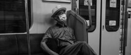 
Fotografia czarno-biała: mężczyzna w maseczce na twarzy siedzący samotnie w wagonie metra.