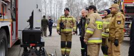 Osoba cywilna mężczyzna funkcjonariusze Państwowej Straży Pożarnej oraz strażacy ochotniczych straży pożarnych stoją obok siebie oglądając sprzęt.