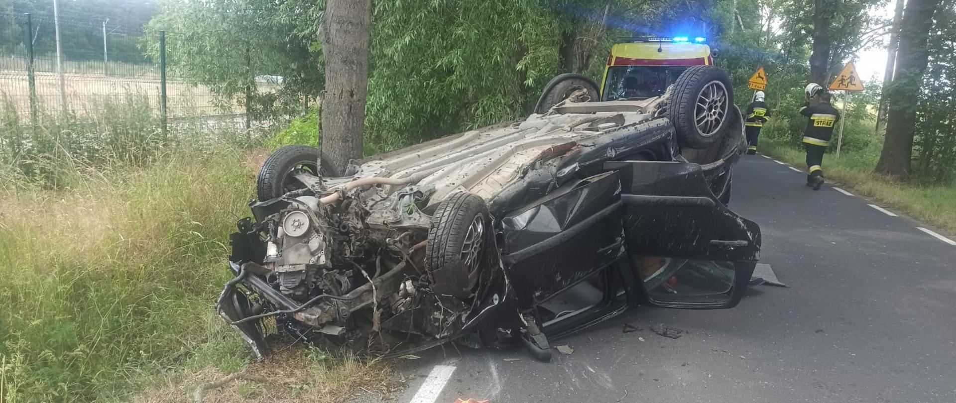 Widać mocno uszkodzony samochód na dachu, leży na drodze