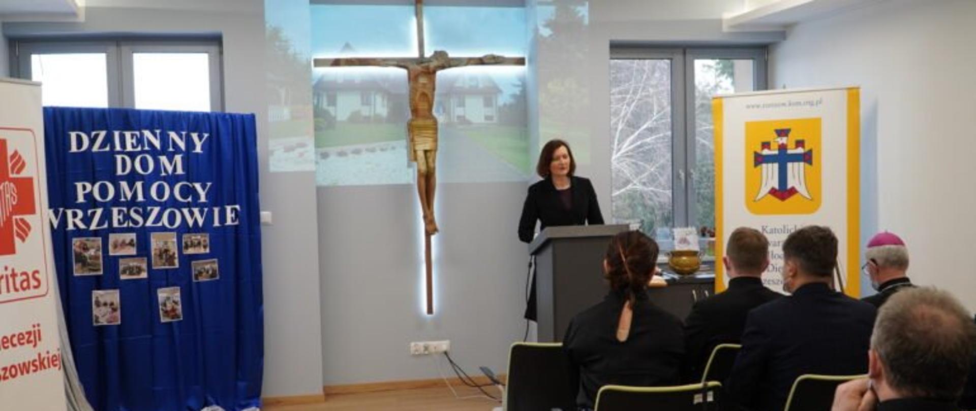 Wojewoda podkarpacki Ewa Leniart przemawia do uczestników uroczystości otwarcia Dziennego Domu Pomocy dla seniorów. Za stojącą przy mównicy wojewodą znajduje się baner Katolickiego Stowarzyszenia Młodzieży, krzyż oraz dekoracja z napisem Dzienny Dom Pomocy w Rzeszowie umieszczonym na niebieskiej kotarze. 