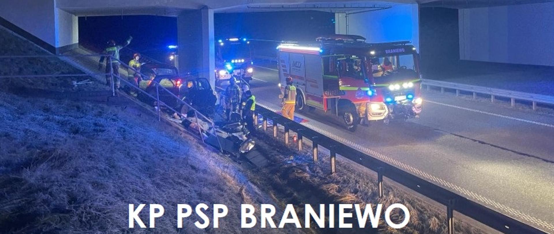 Zdjęcie zrobione nocą. Pod wiaduktem na drodze ustawione trzy samochody strażackie na światłach, oświetlają teren akcji. Z lewej strony uszkodzone auto osobowe, przy nim strażacy, traw akcja ratownicza.