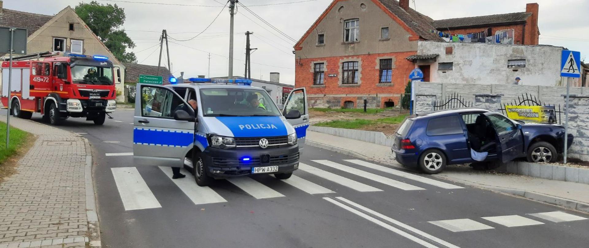 Na fotografii wykonanej na drodze otoczonej budynkami znajdują się: w lewym górnym rogu wóz strażacki OSP Krzęcin, na środku radiowóz policyjny typu bus, który ma otwarte drzwi przy których stoją umundurowani policjanci. Po prawej stronie znajduje się uszkodzony czarny samochód osobowy, które znajduje się w płocie. Tył pojazdu znajduje się na chodniku, a przód pojazdu znajduje się w płocie. Pojazd ma otwarte drzwi od strony pasażera. Radiowóz i samochód osobowy znajdują się na przejściu dla pieszych. W oddali widać drzewo znajdujące się za budynkami. Jest całkowite zachmurzenie.