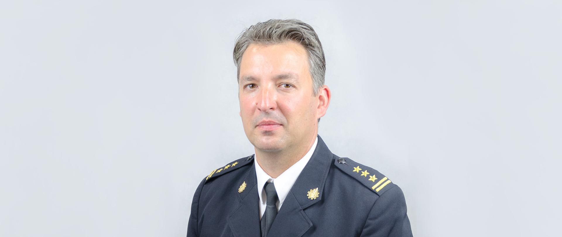 Zdjęcie przedstawia popiersie st. bryg. Roberta Nowakowskiego Komendanta Powiatowego PSP ubranego w mundur wyjściowym na szarym tle.