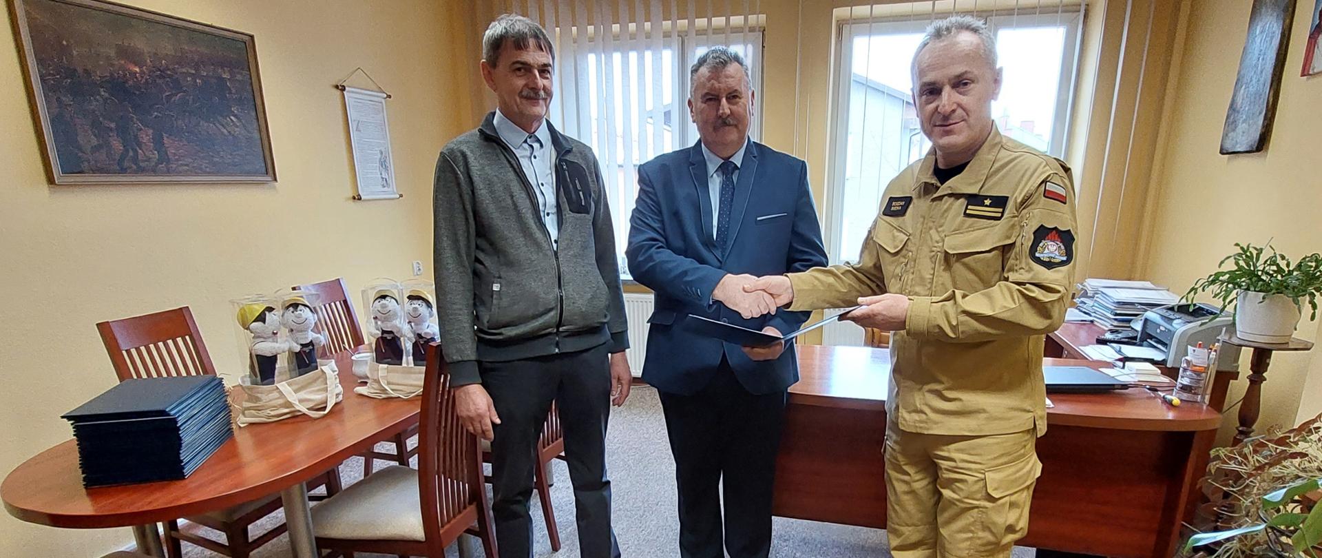 Kolorowa fotografia wykonana w pomieszczeniu, przedstawia spotkanie trzech osób. W pokoju stoją dwie osoby cywilne i jedna w jasnym mundurze służbowym strażaka Państwowej Straży Pożarnej, podczas gratulacji. Z tyłu stoły i krzesła pomieszczenia służbowego. W tle widoczne dwa okna a na jasnych ścianach wiszą obrazy.