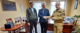 Kolorowa fotografia wykonana w pomieszczeniu, przedstawia spotkanie trzech osób. W pokoju stoją dwie osoby cywilne i jedna w jasnym mundurze służbowym strażaka Państwowej Straży Pożarnej, podczas gratulacji. Z tyłu stoły i krzesła pomieszczenia służbowego. W tle widoczne dwa okna a na jasnych ścianach wiszą obrazy.