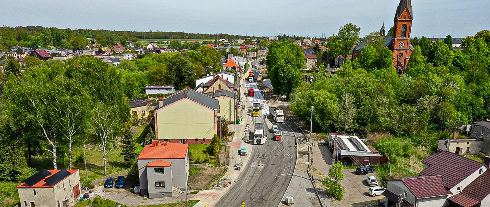 Widok z lotu ptaka na malownicze miasteczko, w którym trwa remont drogi. Na jezdni widać pracujące maszyny budowlane, w tym ciężarówki oraz urządzenia do asfaltowania. Droga jest częściowo pokryta nowym asfaltem, a na jej poboczu ustawiono barierki ochronne. Wzdłuż trasy rozciągają się domy jednorodzinne z czerwonymi dachami oraz budynki gospodarcze. W tle, otoczony drzewami, wyróżnia się kościół z wysoką wieżą. Cała sceneria tonie w zieleni, wskazując na spokojną, urokliwą lokalizację.