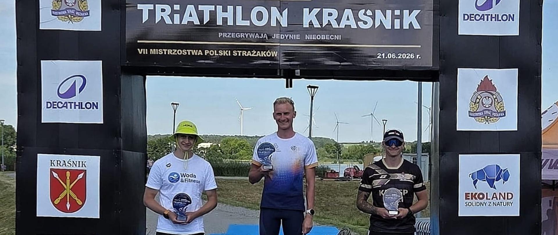 Na podium stoją trzej mężczyźni. Każdy trzyma statuetkę. Za nimi ścianka z napisem "TRIATHLON KRAŚNIK" oraz logo instytucji. 