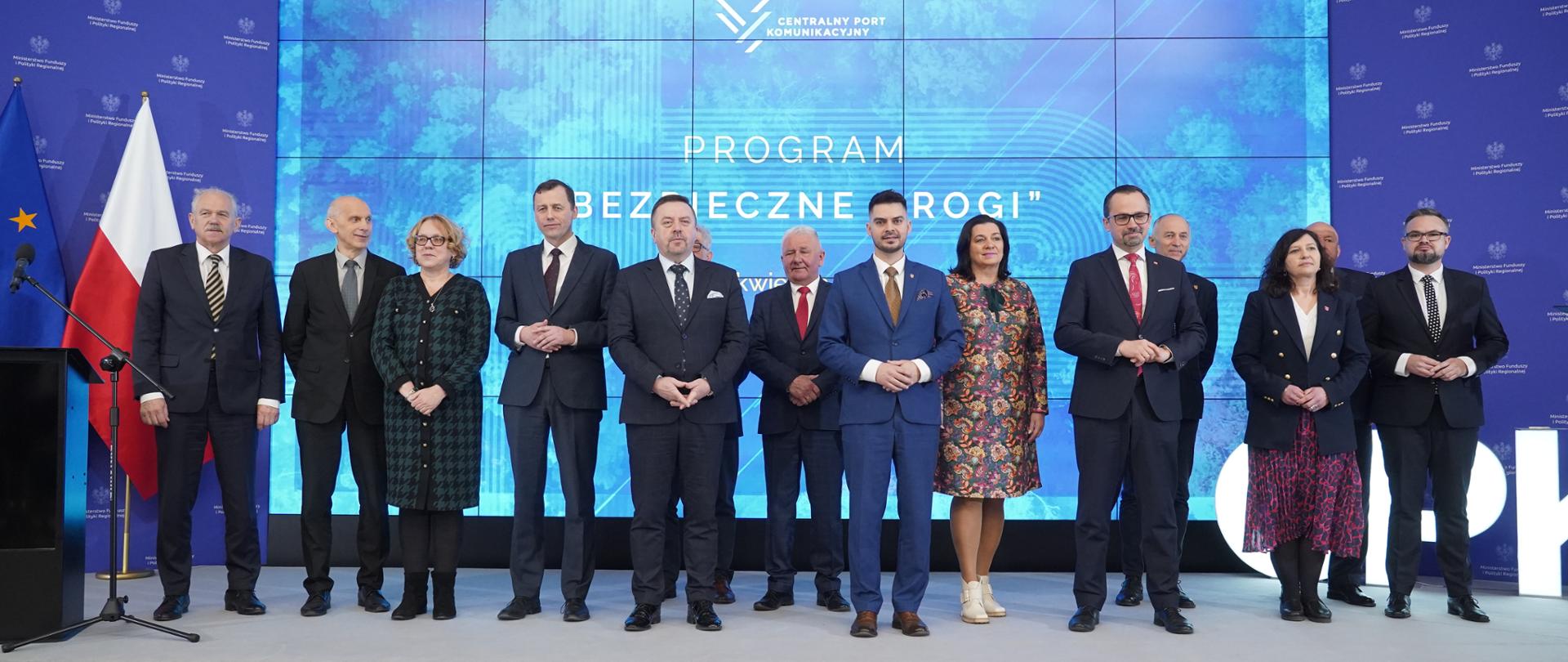 Trzynaście osób stoi obok siebie na scenie. Piąty z prawej stoi wiceminister Marcin Horała.