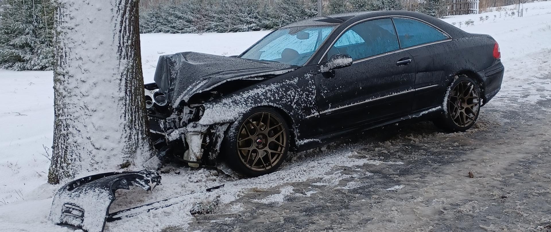 Samochód osobowy uderzył w drzewo. Ma rozbity cały przód, w oddali widoczne są zabudowania. 