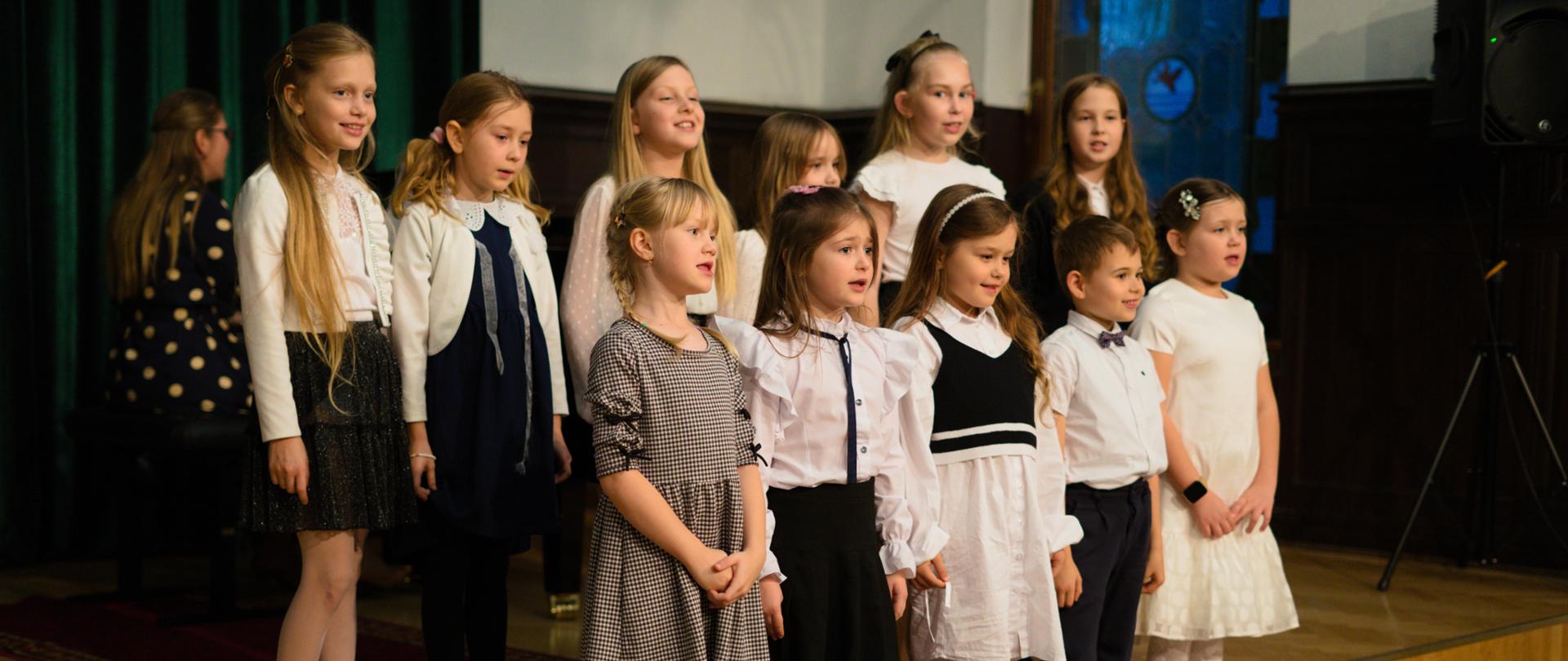 Grupa dzieci w odświętnych strojach śpiewa podczas szkolnej uroczystości na scenie z czerwonym dywanem