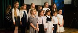 Grupa dzieci w odświętnych strojach śpiewa podczas szkolnej uroczystości na scenie z czerwonym dywanem.