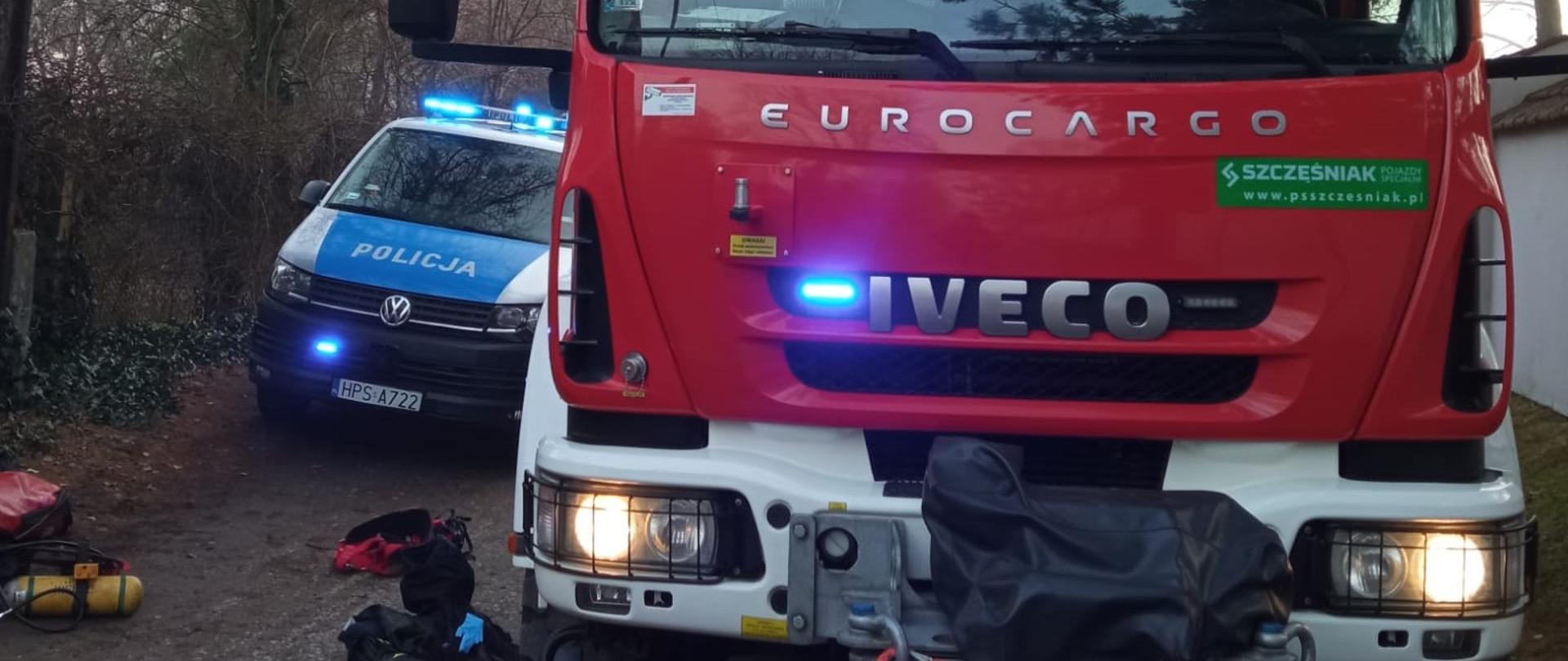 Zdjęcie przedstawia przód samochodu pożarniczego, a po lewej stronie fragment radiowozu policyjnego. Na drodze leżą elementy umundurowania ratownika, który ratował psa.