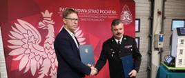 Prezydent Miasta i Komendant Miejski ściskają swoje dłonie. Obaj w rękach trzymają teczki z dokumentami.