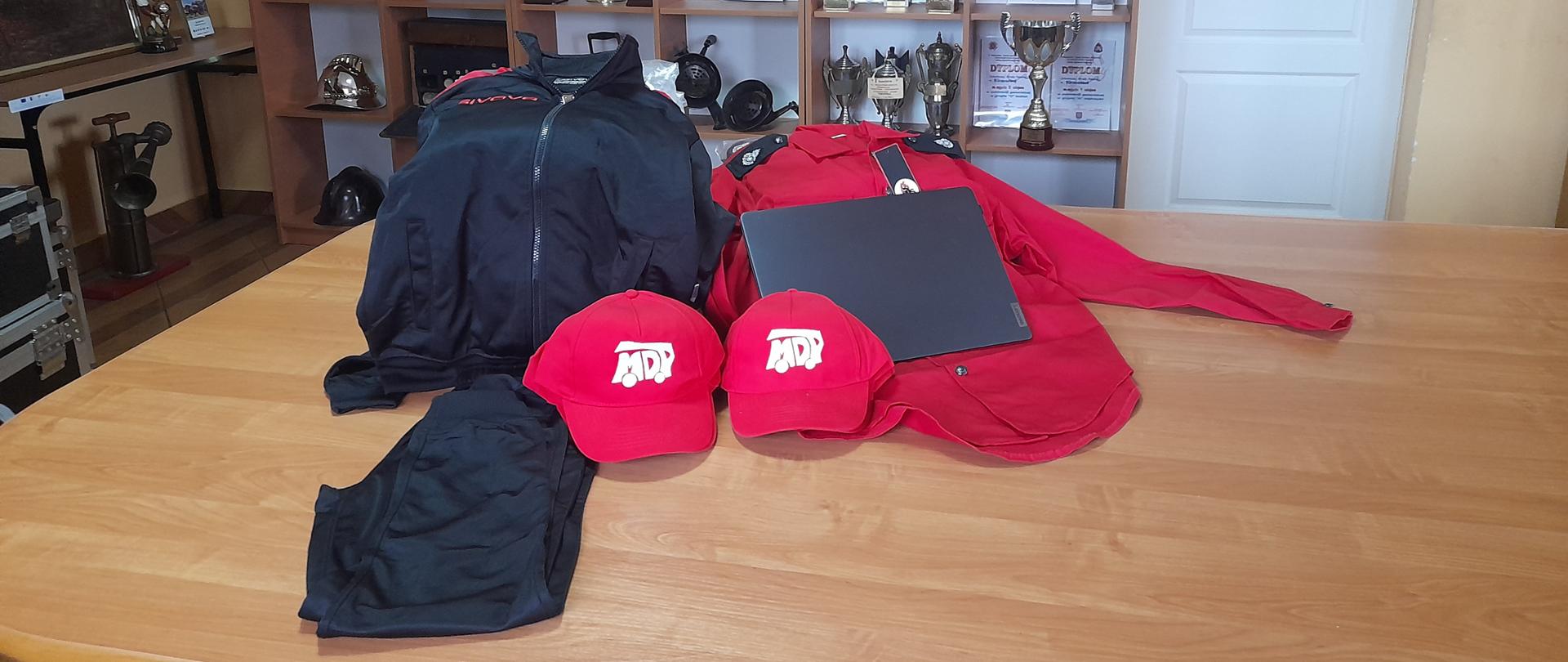 Na brązowym stole na tle szafy z pucharami leżą czerwone i granowe ubrania, czapki i laptop.