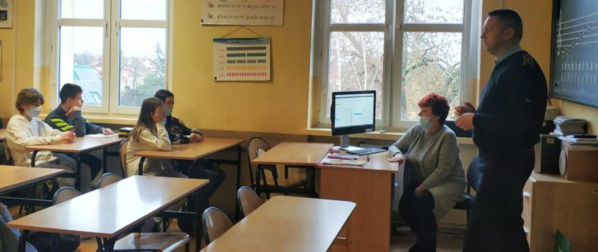 Zdjęcie przedstawia strażaka prowadzącego zajęcia z dziećmi w szkole. Dzieci siedzą przy ławkach. Strażak w umundurowaniu służbowym stoi przy tablicy. Obok niego przy biurku siedzi Pani Nauczycielka. Zdjęcie zrobione od strony wejścia do sali