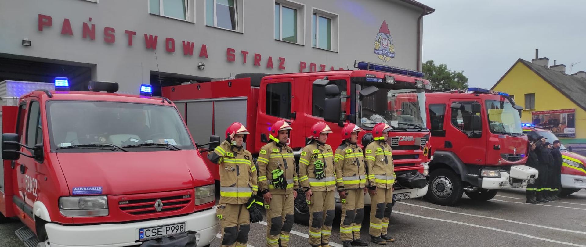 Zdjęcie wykonano porą dzienną, strażacy Państwowej Straży Pożarnej ubrani w mundury specjalne oraz hełmy oraz strażacy Ochotniczej Straży Pożarnej w Sępólnie Krajeńskim ubrani w mundury koszarowe stoją przed budynkiem Komendy Państwowej Straży Pożarnej w Sępólnie Krajeńskim na baczność. Za nimi samochody pożarnicze, oraz otwarte bramy garażowe budynku komendy.