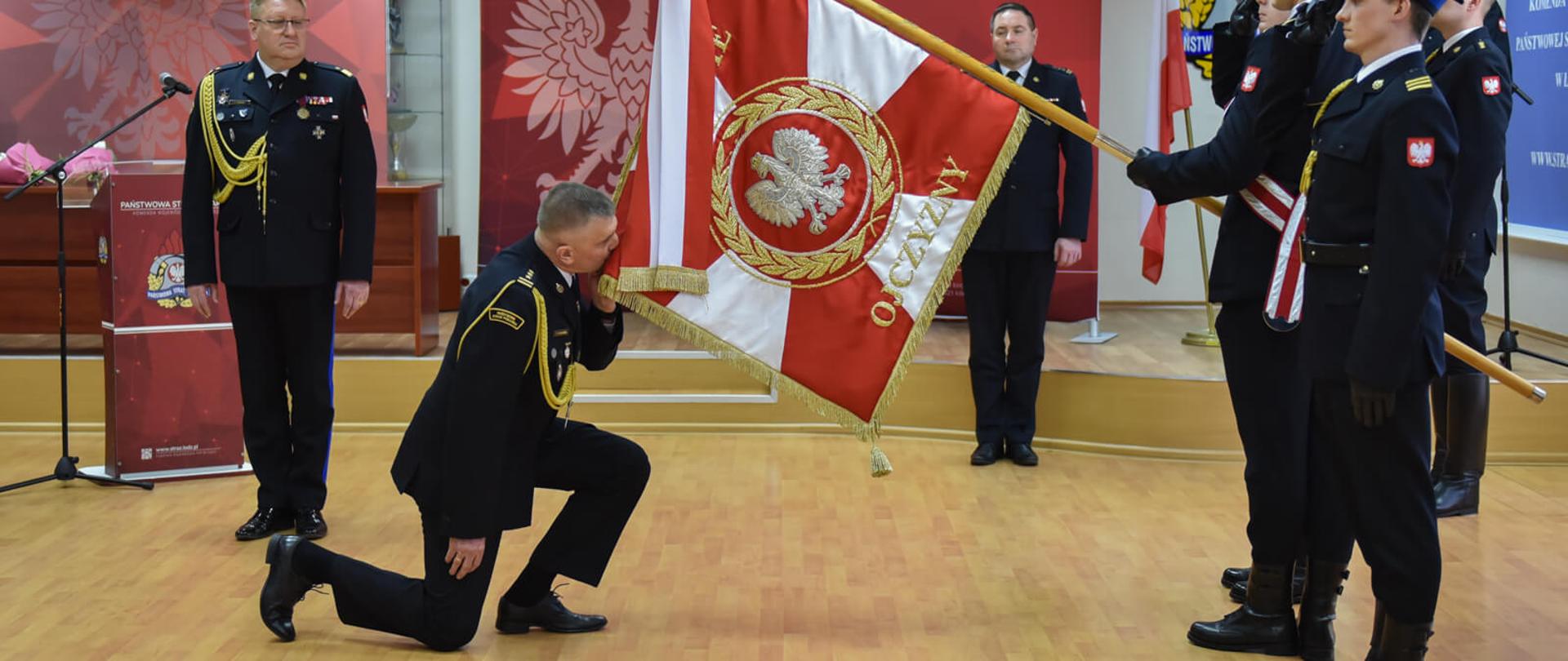 starszy brygadier Paweł Malinowski przyklęka i całuje sztandar komendy, na przeciwko stoi poczet sztandarowy