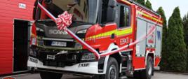 Nowy, czerwony wóz strażacki marki Scania z numerem bocznym "639[M]31" stoi przed budynkiem z czerwonymi drzwiami garażowymi. Wóz jest udekorowany biało-czerwonymi girlandami i dużą biało-czerwoną kokardą na przodzie. Na budynku strażnicy powiewa polska flaga. Na boku wozu widnieje logo OSP Rybno. W tle widać zielone drzewa.