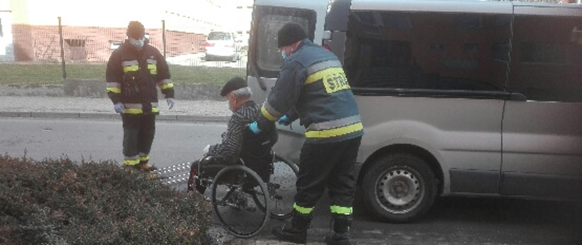 Obraz przedstawia strażaków pomagających osobie na wózku dostać się do samochodu, którym zostanie przetransportowany na szczepienie.