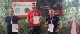 Podium zawodów, na którym stoi trzech mężczyzn z medalami na szyji oraz pucharami i dyplomami w dłoniach.