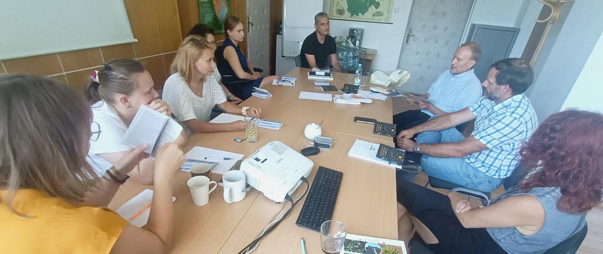 Przy stole siedzi grupa kilku kobiet i trzech mężczyzn. Na stole leżą książki. Jeden z mężczyzn mówi.