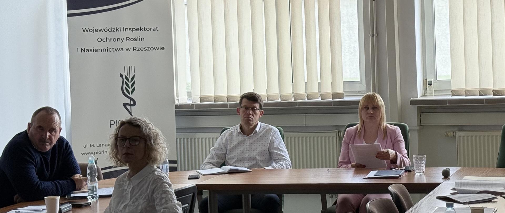 Kobieta i mężczyzna siedza przy stole w jasnym pomieszczeniu. Za nimi są okna. Po lewej stronie stoi biało granatowy baner z logiem instytucji koło którego siedzi mężczyzna i kobieta.