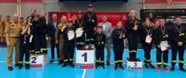 
Uczestnicy II Halowych Młodzieżowych Zawodów Sportowo-Pożarniczych pozują na podium z medalami, pucharami i dyplomami po zakończonej rywalizacji.