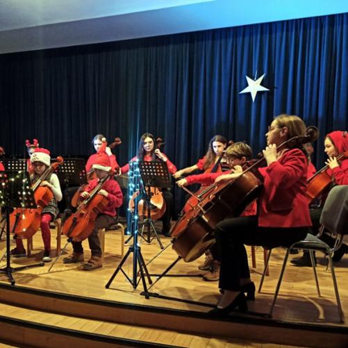 Zdjęcie przedstawia grupę dzieci, grających na wiolonczelach, w pozycji siedzącej - na krzesłach. Orkiestra wiolonczelowa siedzi na drewnianej scenie. W tle widać granatową kotarę z zawieszoną , świecącą gwiazdą. Wszyscy ubrani są w świąteczne, czerwone stroje.