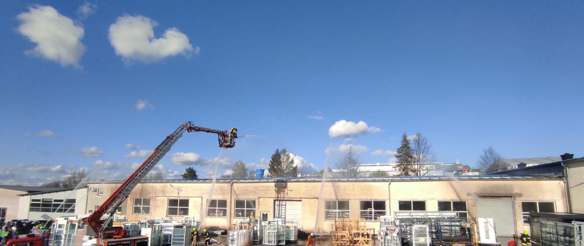 Na zdjęciu po prawej stronie widać rozstawiony samochód pożarniczy z koszem z którego podawana jest woda na dach budynku. Po prawej stronie widać strażaków podających również wodę na dach i ściany budynku