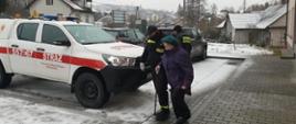 Strażak prowadzi starszą kobietę. Po jego prawej samochód strażacki koloru białego.