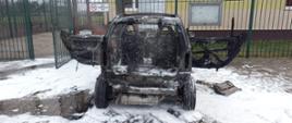 Pożar smarta. Parking przy Orliku w Bojanowie. Na pierwszym planie spalony pojazd. W tle ogrodzenie posesji oraz budynek. Wokół samochodu pozostałości piany gaśniczej. 