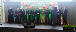 grupa osób stoi na scenie, jedna z nich przemawia do mikrofonu, niektórzy biją brawa, w tle wisi baner z napisem Wystawa Wielkanocna Glinojeck, na którym widoczne są kurze jajka