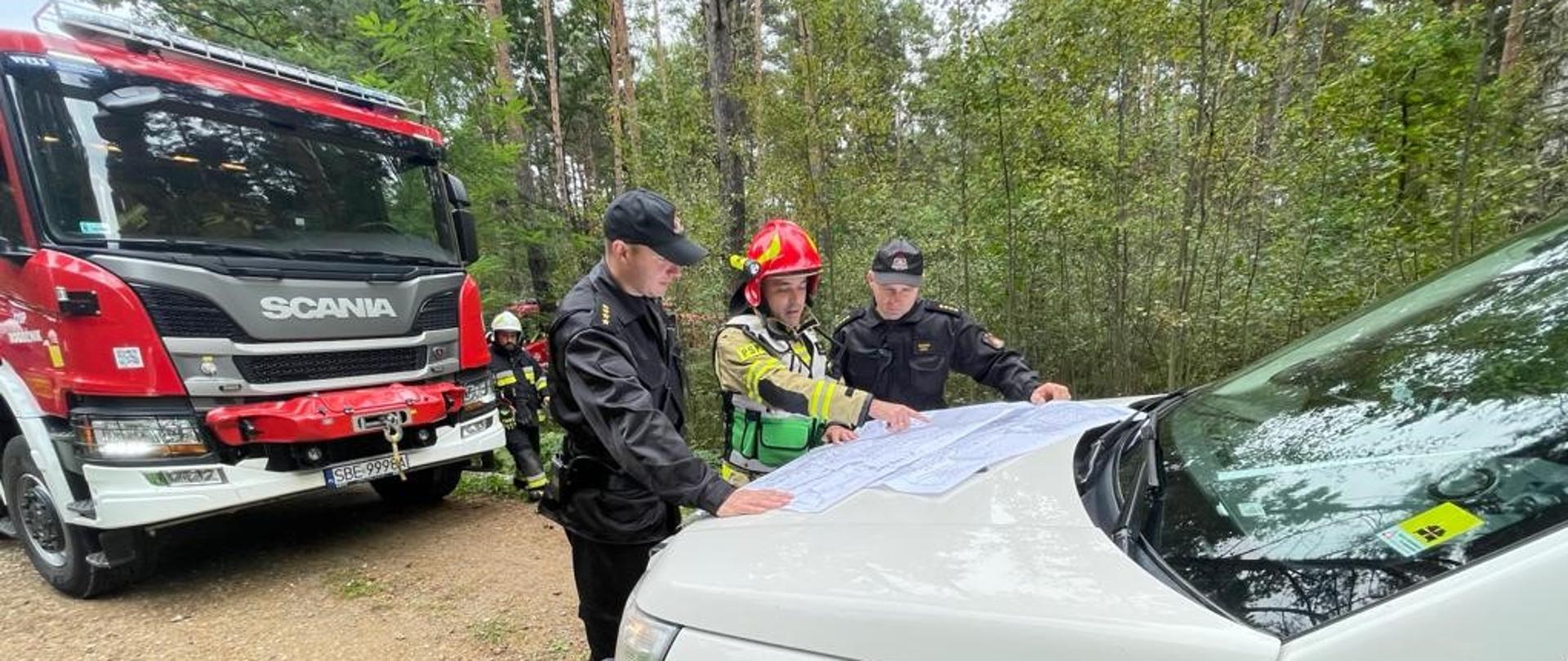 Zdjęcie zrobione na jednej z dróg pożarowych w Leśnictwie Wymysłów, przedstawia dowódcę Kompanii Odwodowej nr 1 "Będzin", który pokazuje na mapie znajdującej się na masce jednego z samochodów zakres prowadzonych działań dwóm pozostałym uczestnikom ćwiczeń.
