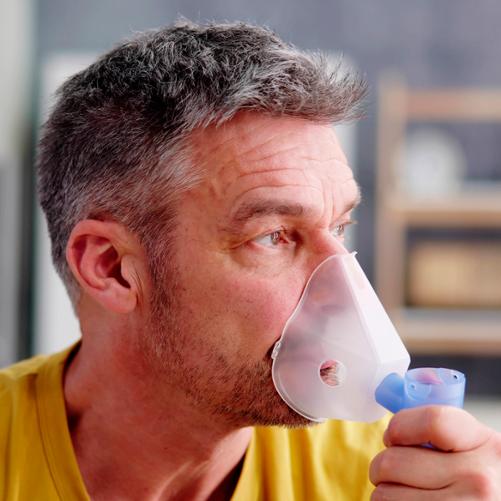 Mężczyzna z lelkko siwiejącymi włosami w żółtej koszulce przykłada do twarzy maskę nebulizatora.