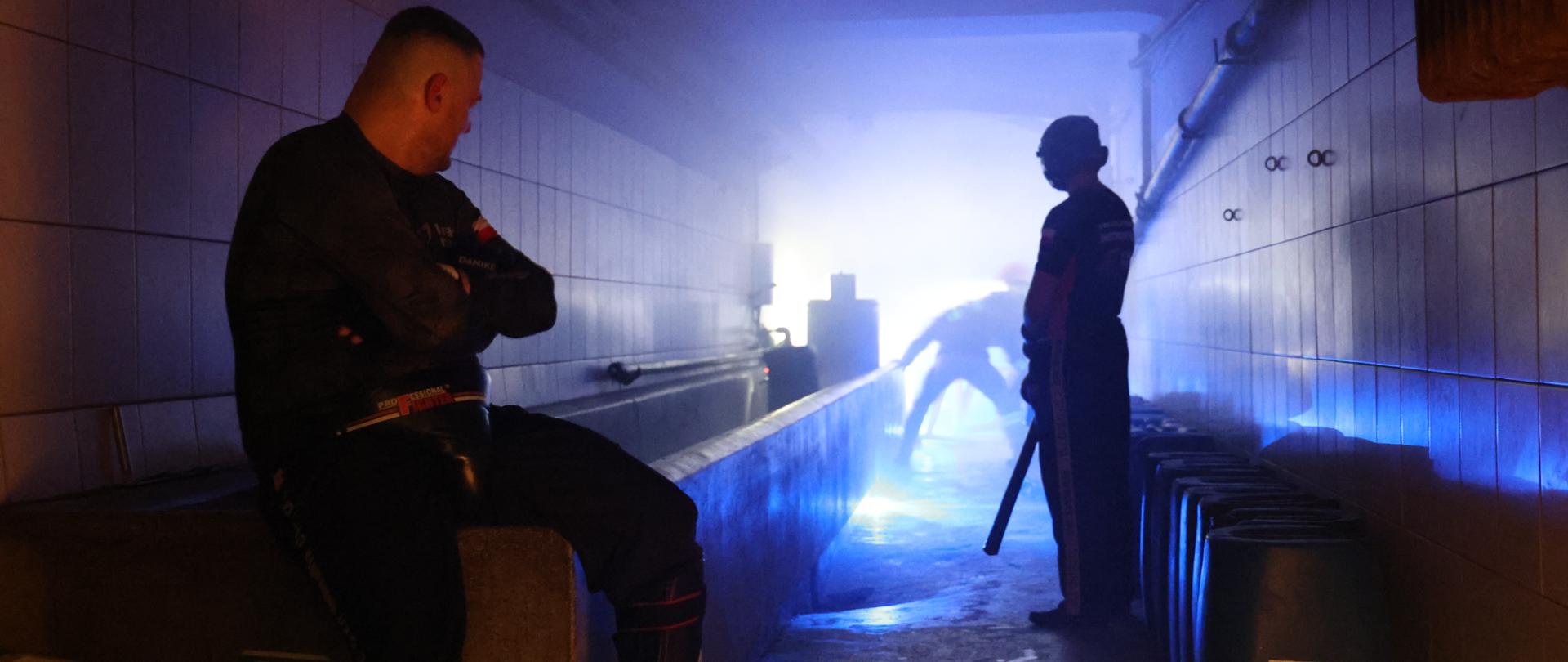 Zdjęcie zrobione w ciemnej zadymionej piwnicy. Na pierwszym planie widoczni dwaj mężczyźni. Osoby walczą ze sobą co jest częścią przeprowadzanych warsztatów