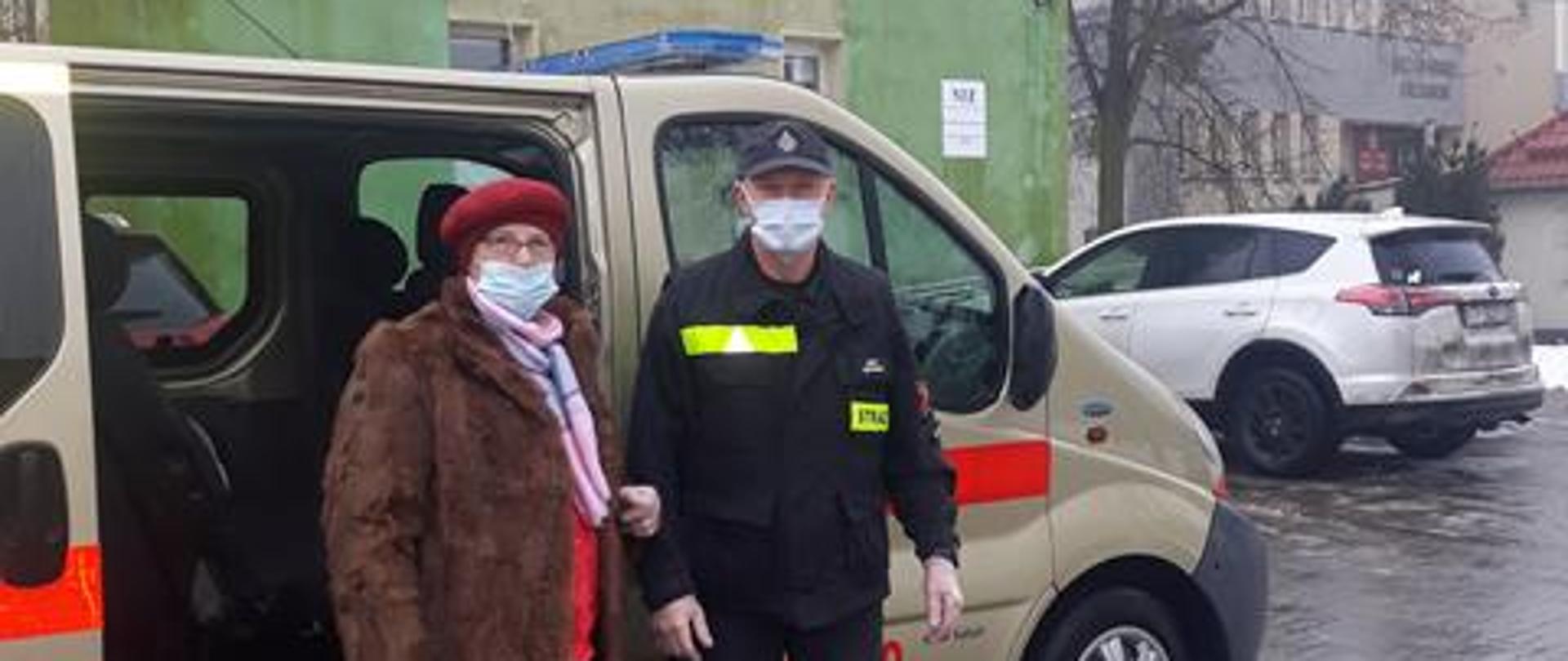 Na zdjęciu widać kobietę i strażaka po wyjściu z pojazdu OSP Elgiszewo. Kobieta przy pomocy strażaka udaje się do punktu szczepień.