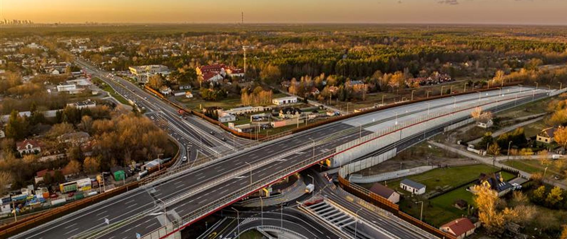 Na zdjęciu widać fragment infrastruktury drogowej związanej z trasą S17 w rejonie Warszawy Wschodniej.
Zdjęcie przedstawia odcinek drogi ekspresowej lub węzła komunikacyjnego w fazie eksploatacji lub dokumentacji.
Elementy widoczne na pierwszym planie, to jezdnia asfaltowa z wyraźnym oznakowaniem poziomym (linie rozdzielające pasy ruchu).
Barierki energochłonne wzdłuż krawędzi drogi. Na poboczu widoczne są słupki kilometrowe lub oznaczenia techniczne. W oddali widoczne są elementy infrastruktury – wiadukt lub estakada nad drogą.
Roślinność wzdłuż drogi, głównie trawy i krzewy, a dalej zabudowa lub obiekty techniczne. Dominują odcienie szarości (nawierzchnia), zieleni (roślinność) i niebieskiego (niebo).
Pogoda wydaje się dobra, niebo jest jasne z lekkim zachmurzeniem.