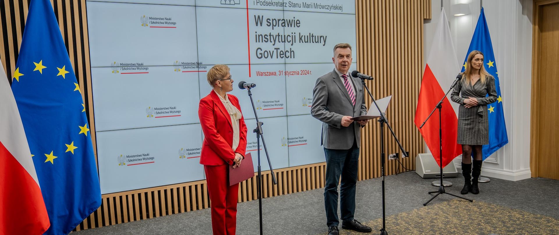 Na sali stoi minister Wieczorek i wiceminister Mrówczyńska w czerwonym garniturze, minister mówi do mikrofonu na stojaku.